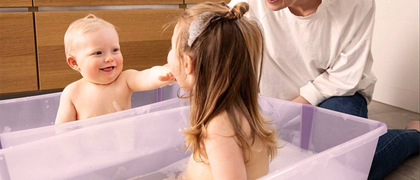 Zwei Kinder haben Spaß beim Baden, jedes in einer eigenen Wanne, während eine Betreuerin in der Nähe lächelt – alles im Badezimmer.
