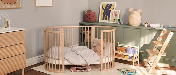 Gemütliches Kinderzimmer mit einem runden Holzkinderbett, Stofftieren, einem kleinen Bücherregal, einem hölzernen Hochstuhl und pastellfarbener Dekoration auf einem grünen Regal.