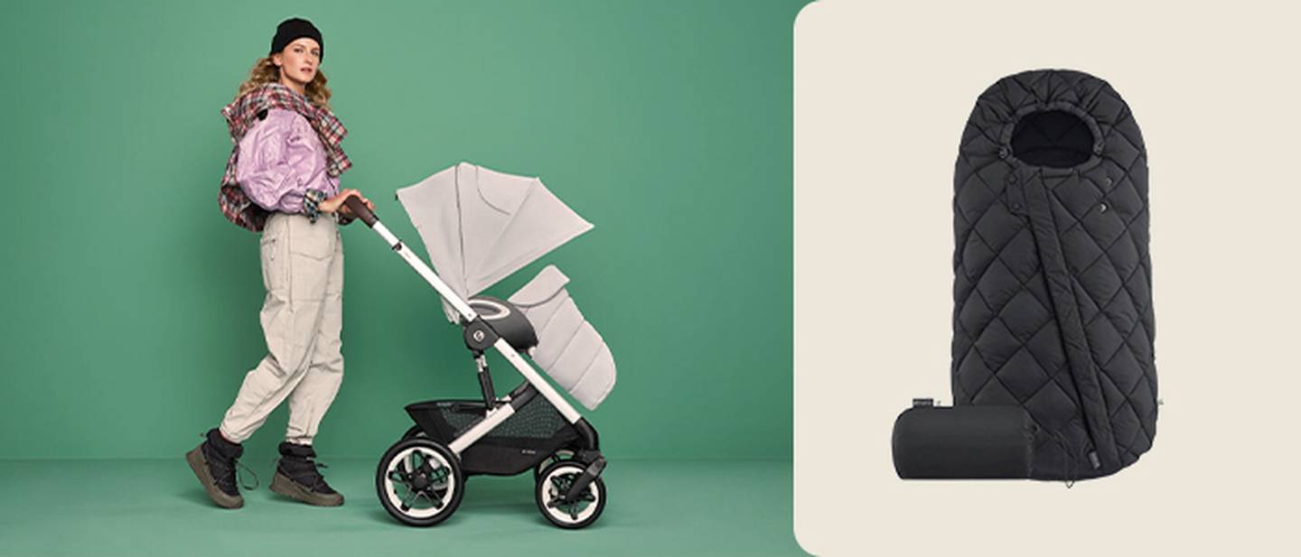 Eine Frau schiebt einen rosa Kinderwagen vor grünem Hintergrund; im rechten Bildausschnitt sind ein schwarzer, gesteppter Fußsack und ein zusammengerolltes Zubehörteil zu sehen.