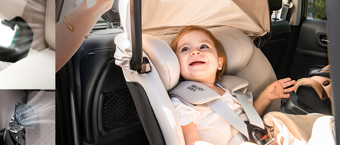 Ein glückliches Kind in einem Autositz mit beigem Bezug, geschützt durch ein Verdeck. Das Kind blickt nach oben, lächelt und streckt den Arm nach vorne.