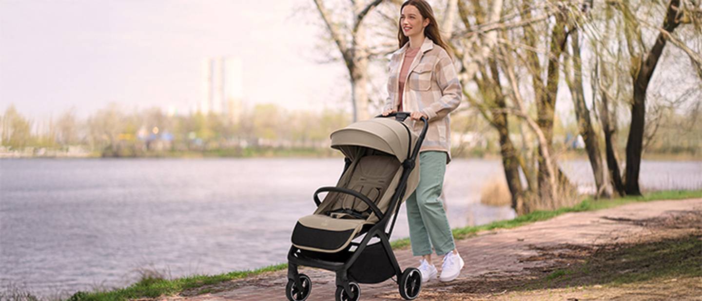 Eine Frau schiebt einen beigen Kinderwagen auf einem Uferweg entlang, umgeben von Bäumen und einer ruhigen Wasserlandschaft im Hintergrund.