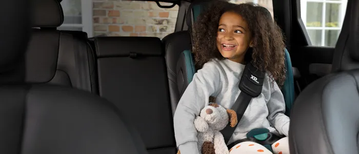 Ein Kind lächelt im Kindersitz, hält einen Stoffhund im Arm und trägt einen grauen Pullover sowie eine Leggings mit Punkten.