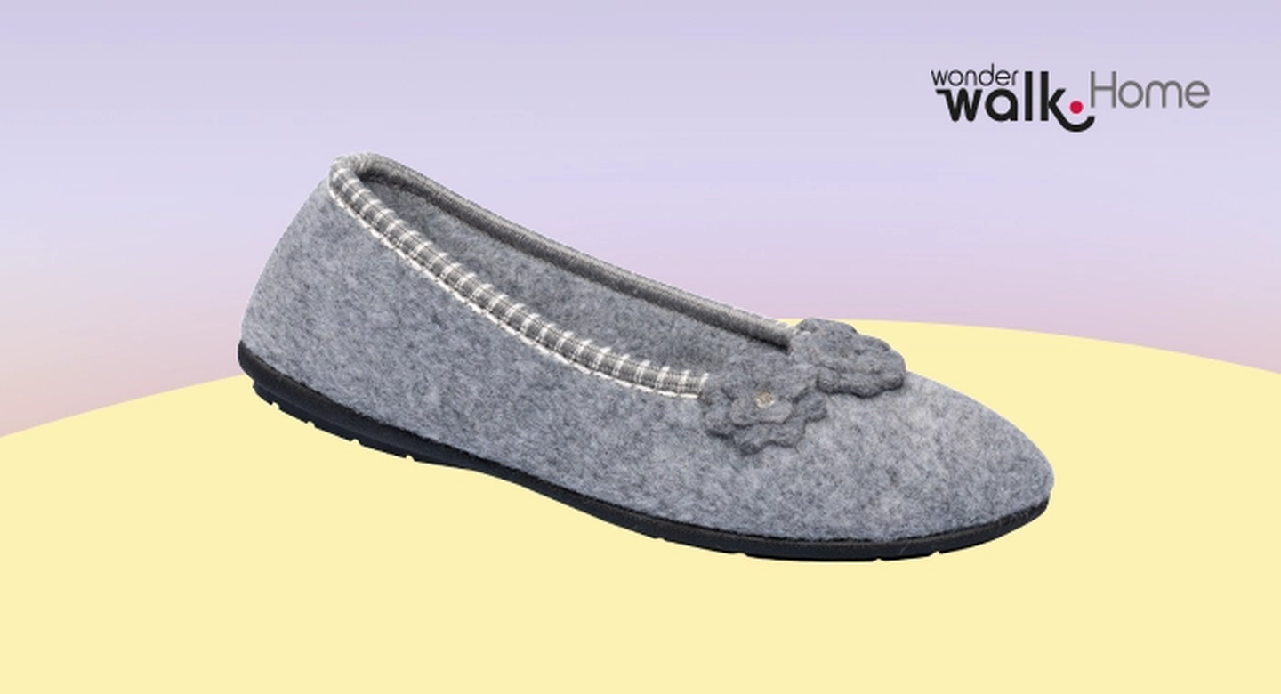 Grauer Hausschuh mit Blumenmuster und schwarzer Sohle auf zartem Pastellhintergrund. Logo „Wonder Walk Home“ in der oberen Ecke.