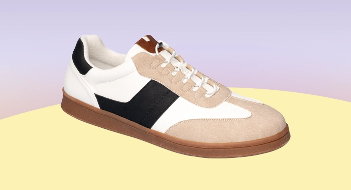 Lässiger Sneaker mit beigen, weißen und schwarzen Einsätzen, brauner Sohle und weißen Schnürsenkeln vor pastellfarbenem Hintergrund.