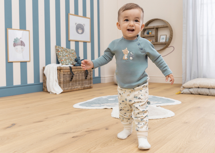 Ein lächelndes Kleinkind, das in einem gestreiften Kinderzimmer auf einem Holzboden laufen lernt, trägt einen türkisfarbenen Pullover und eine gemusterte Hose.