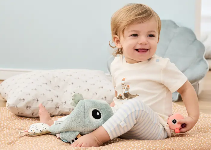 Ein lächelndes Kleinkind sitzt auf einer Decke, hält ein Plüschtier im Arm, umgeben von einem Kissen und weiteren Kuscheltieren. Es trägt ein weißes Hemd und eine gestreifte Hose.