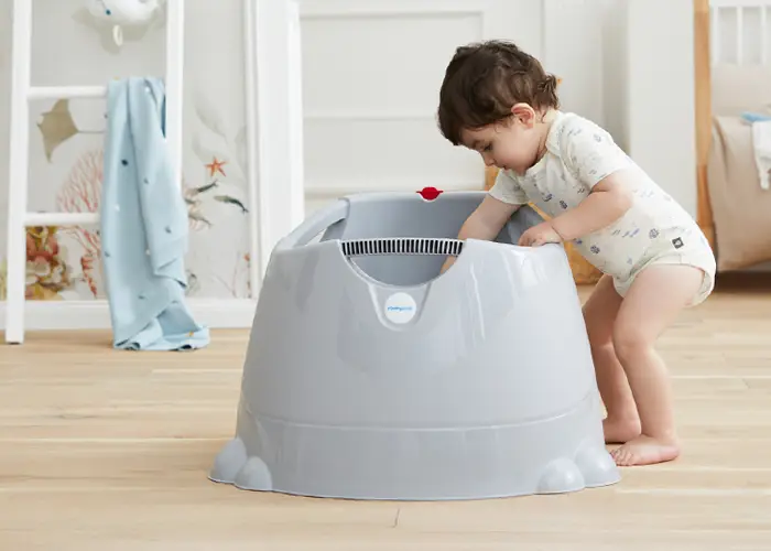 Kleinkind im Strampler lehnt sich an eine große graue Plastik-Babytöpfchen-/Trainingswanne auf dem Holzboden des Kinderzimmers.