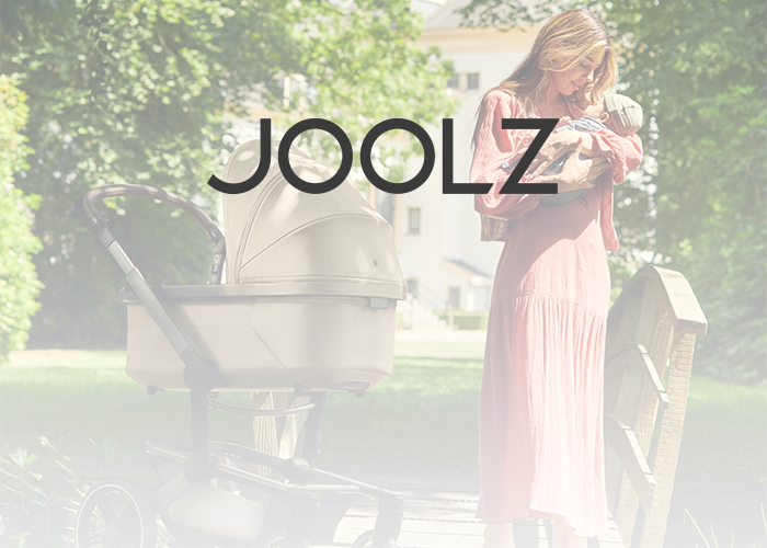 Frau in einem rosa Kleid hält ein Baby neben einem Kinderwagen auf einer Holzbrücke, darüber das "Joolz"-Logo.