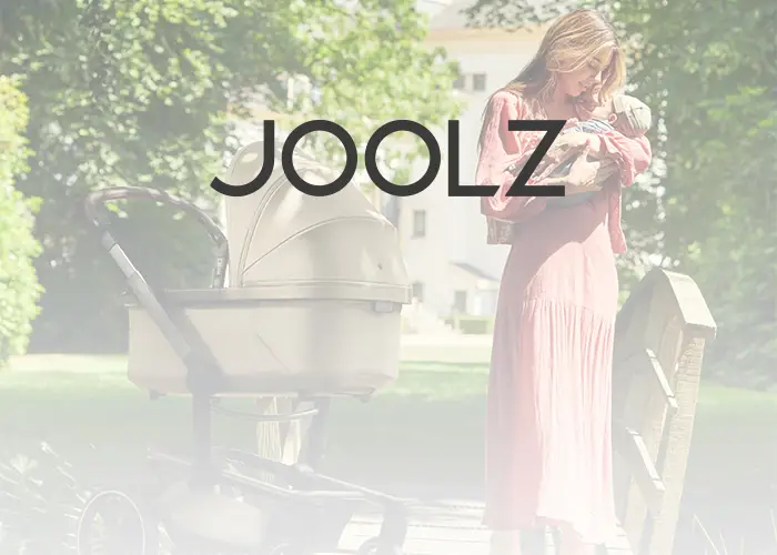 Frau in einem rosa Kleid hält ein Baby neben einem Kinderwagen auf einer Holzbrücke, darüber das "Joolz"-Logo.
