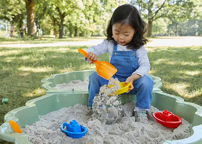 Ein junges Mädchen spielt im Sandkasten und füllt mit einer gelben Schaufel einen grünen Eimer mit Sand. Bunte Spielzeugboote liegen in der Nähe.