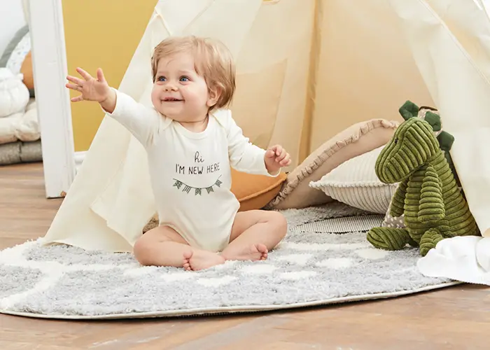 Ein lächelndes Baby in einem weißen Strampler sitzt auf einem grauen Teppich und streckt die Arme aus. In der Nähe stehen ein grüner Plüsch-Dinosaurier und Kissen neben einem kleinen Zelt.