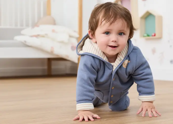 Ein Baby in einem blauen Kapuzenstrampler krabbelt lächelnd auf einem Holzboden in einem gemütlichen, schön eingerichteten Kinderzimmer mit Kinderbett und Dekoration.