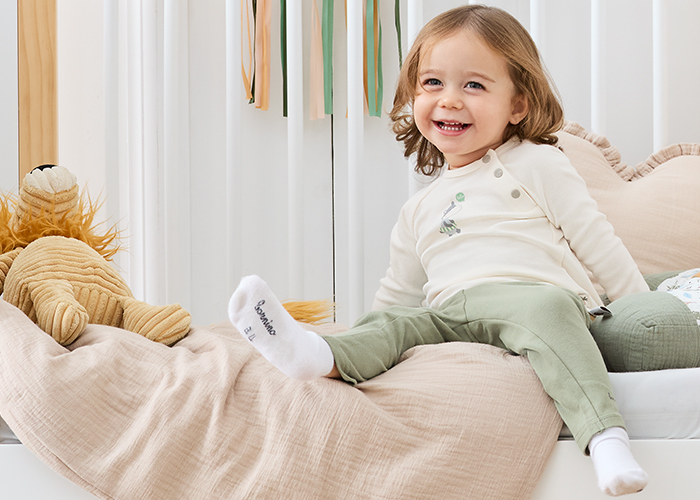 Ein lächelndes Kleinkind in einem weißen Oberteil und einer grünen Hose sitzt auf einem Bett mit einer beigen Decke und einem Spielzeuglöwen in der Nähe; im Hintergrund ist eine bunte Dekoration zu sehen.