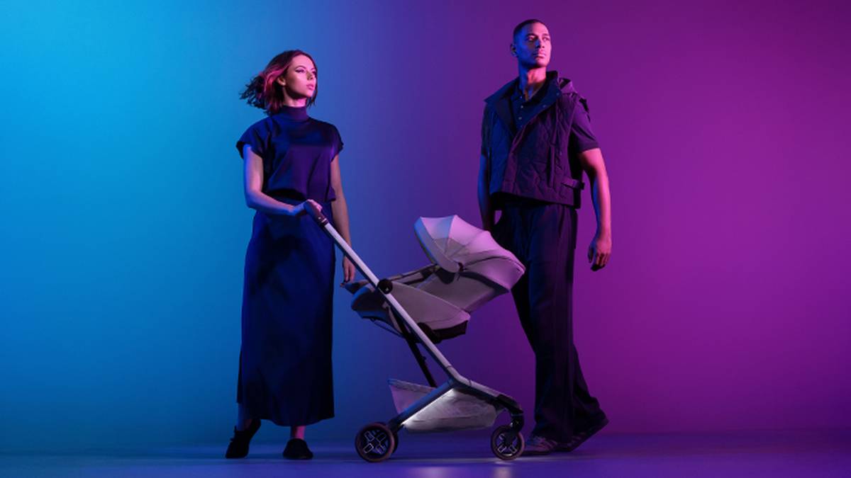 Eine Frau in einem langen Kleid und ein Mann in einer Weste stehen unter blauer und rosa Beleuchtung; die Frau schiebt einen modernen Kinderwagen.