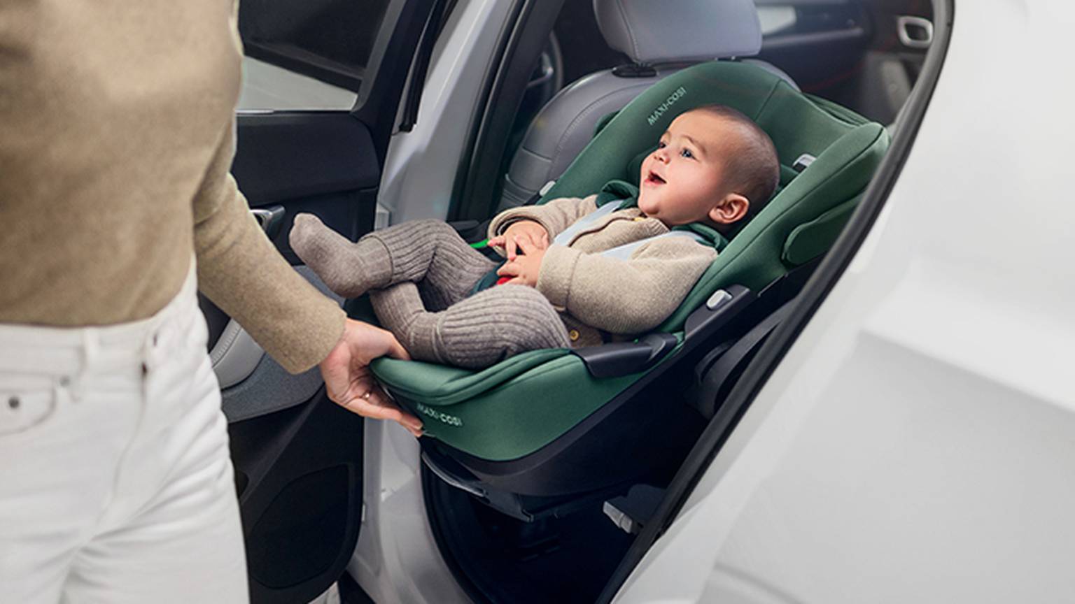 Ein Baby in einem grünen Kindersitz lächelt einen Erwachsenen an, der neben der offenen Autotür steht. Der Erwachsene trägt einen beigen Pullover und eine weiße Hose.