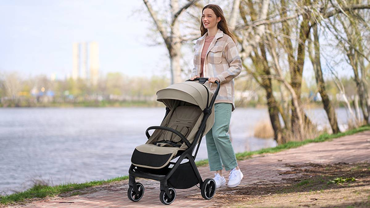 Eine Frau geht an einem Uferweg entlang und schiebt einen leeren beigen Kinderwagen. Sie ist leger gekleidet mit einer karierten Jacke und einer grünen Hose.