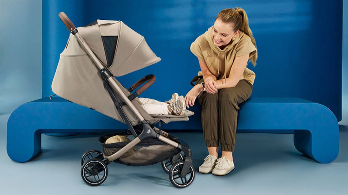 Eine Frau in Freizeitkleidung lächelt ein Baby in einem beigen Kinderwagen an. Sie befinden sich in einem modernen Raum mit einer blauen Bank und blauem Hintergrund.