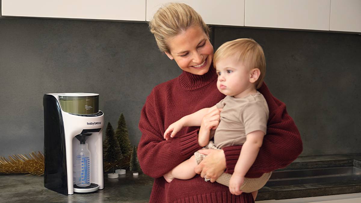Eine Frau in einem roten Pullover lächelt, während sie ein kleines Kind in einer Küche im Arm hält. Neben ihr auf der Arbeitsfläche steht ein Säuglingsnahrungsautomat.