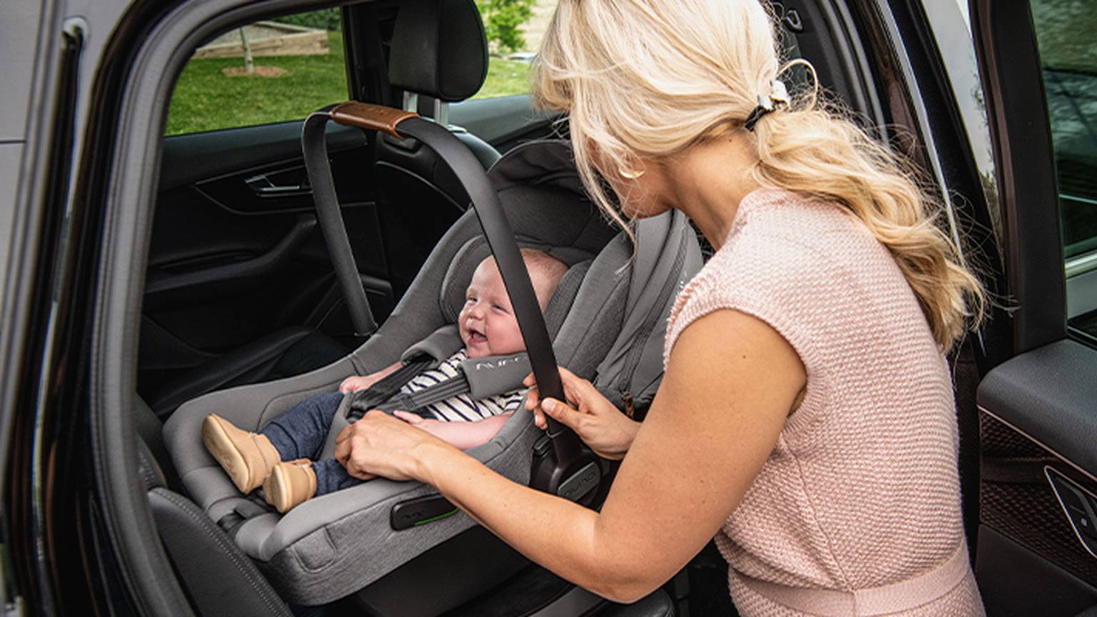 Frau sichert ein lächelndes Baby in einem Kindersitz in einem Fahrzeug bei geöffneter Autotür.
