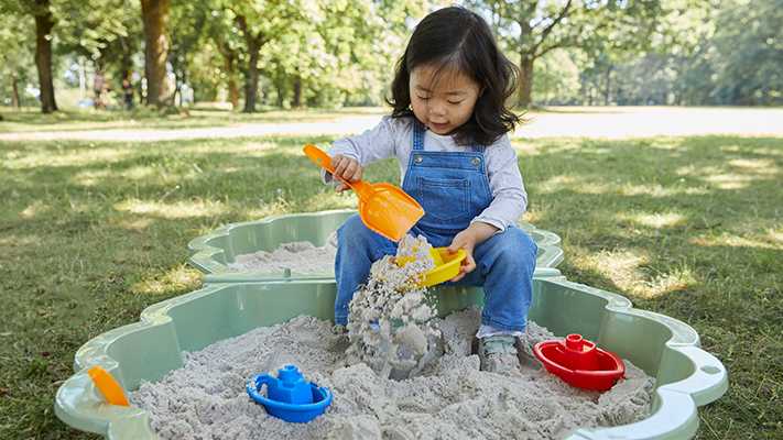 Ein Kind in blauer Latzhose spielt draußen in einem Sandkasten mit einer gelben Schaufel und einem roten Eimer, umgeben von Spielzeugbooten.