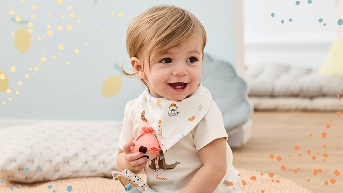 Ein lächelndes Kleinkind sitzt auf einer weichen Matte, trägt ein bedrucktes Lätzchen und hält ein kleines Spielzeug in der Hand. Das Zimmer ist mit fröhlichen Mustern dekoriert.
