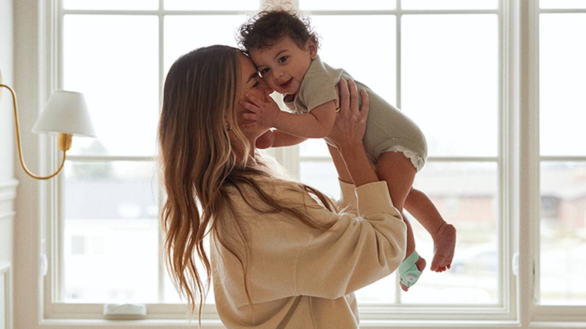 Eine Frau in einem beigen Pullover hebt ein lächelndes Baby im Strampler in der Nähe eines hellen Fensters hoch und schafft so einen warmen, freudigen Moment.
