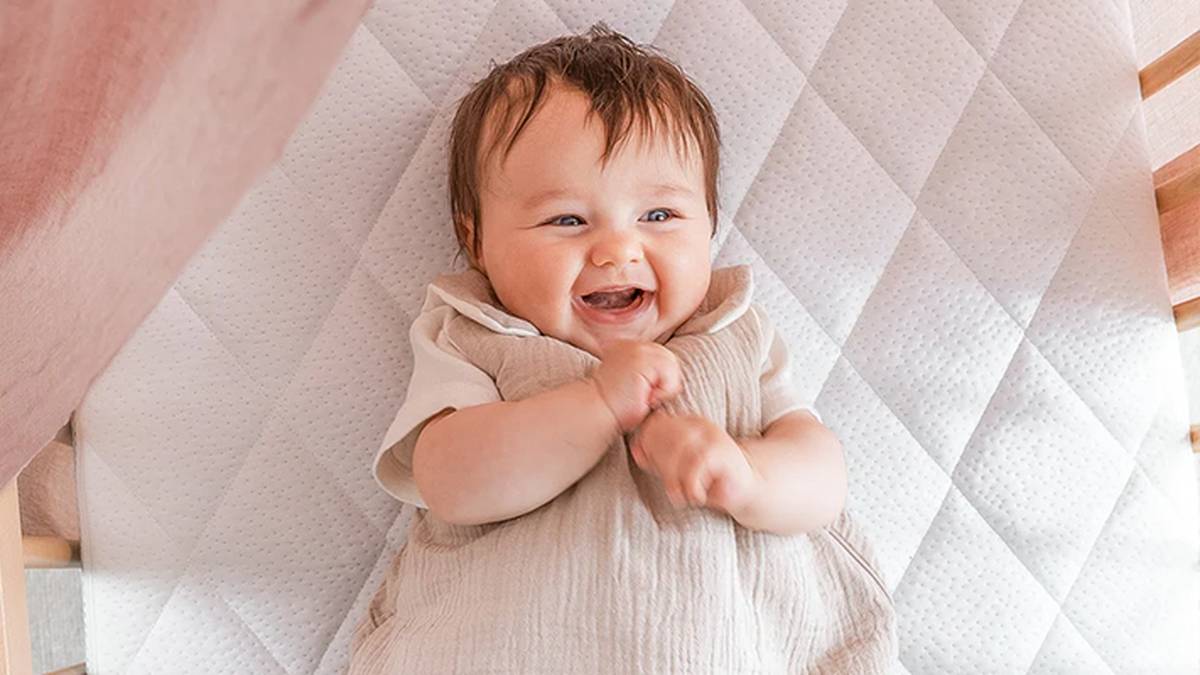 Ein lächelndes Baby liegt auf einer gesteppten Matratze, trägt ein helles Outfit und hat die Hände vor der Brust verschränkt.