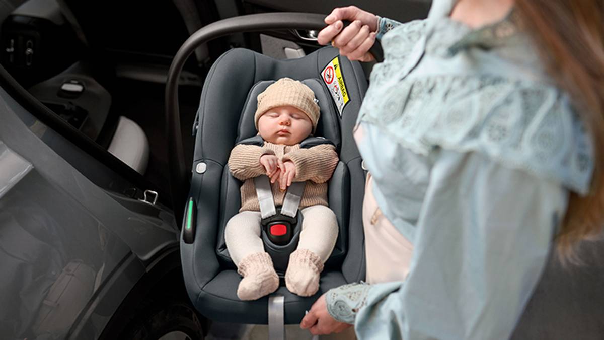 Ein Baby schläft in einem Autositz und trägt beigefarbene Kleidung und ein passendes Mützchen. Ein Erwachsener trägt den Sitz in der Nähe einer Autotür.
