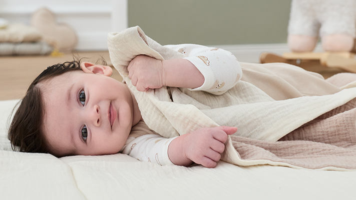 Ein Baby liegt zufrieden auf einer weichen Unterlage, hält eine helle Decke und trägt ein weißes Outfit mit aufgedruckten Mustern.