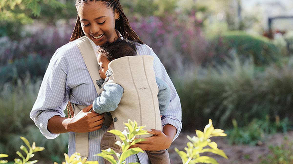 Eine Frau lächelt, während sie ein Baby in einer beigen Trage hält, umgeben von Grün und weichgezeichneten Pflanzen im Hintergrund.