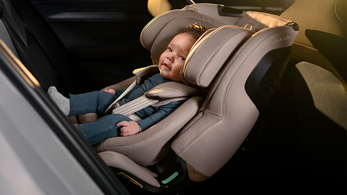 Ein Baby sitzt sicher in einem beigen Kindersitz, sieht zufrieden aus und trägt ein blaues Outfit, während es sich in einem Auto befindet.