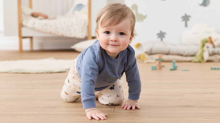 Ein lächelndes Baby in einem blauen Hemd und einer gemusterten Hose krabbelt auf einem Holzboden in einem gemütlichen Zimmer; im Hintergrund sind Spielzeug und ein Bett zu sehen.