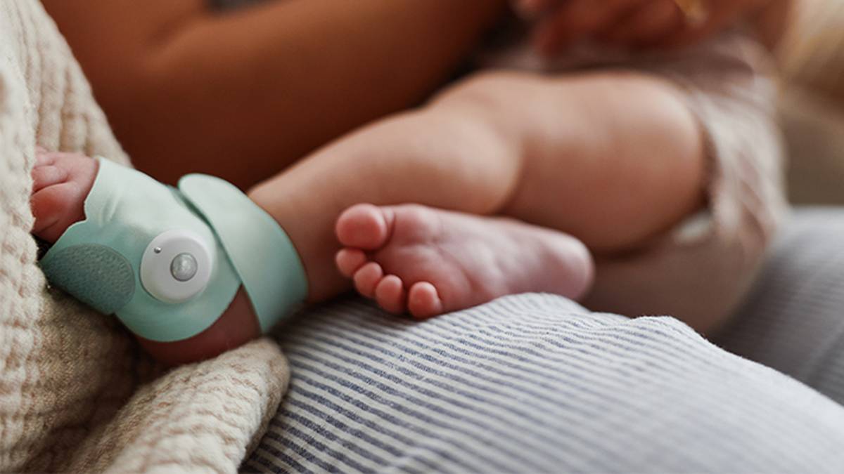Der Fuß eines Babys, an dem ein mintgrüner Monitor angebracht ist, ruht auf einem gestreiften Kissen; im Hintergrund ist der Arm eines Erwachsenen zu sehen.