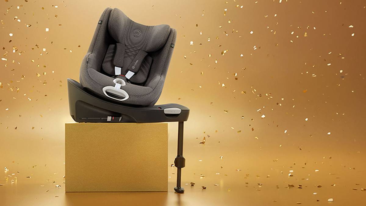 Ein grauer Babysitz auf einer goldenen Schachtel, umgeben von herabfallendem Konfetti, vor einem goldenen Hintergrund.