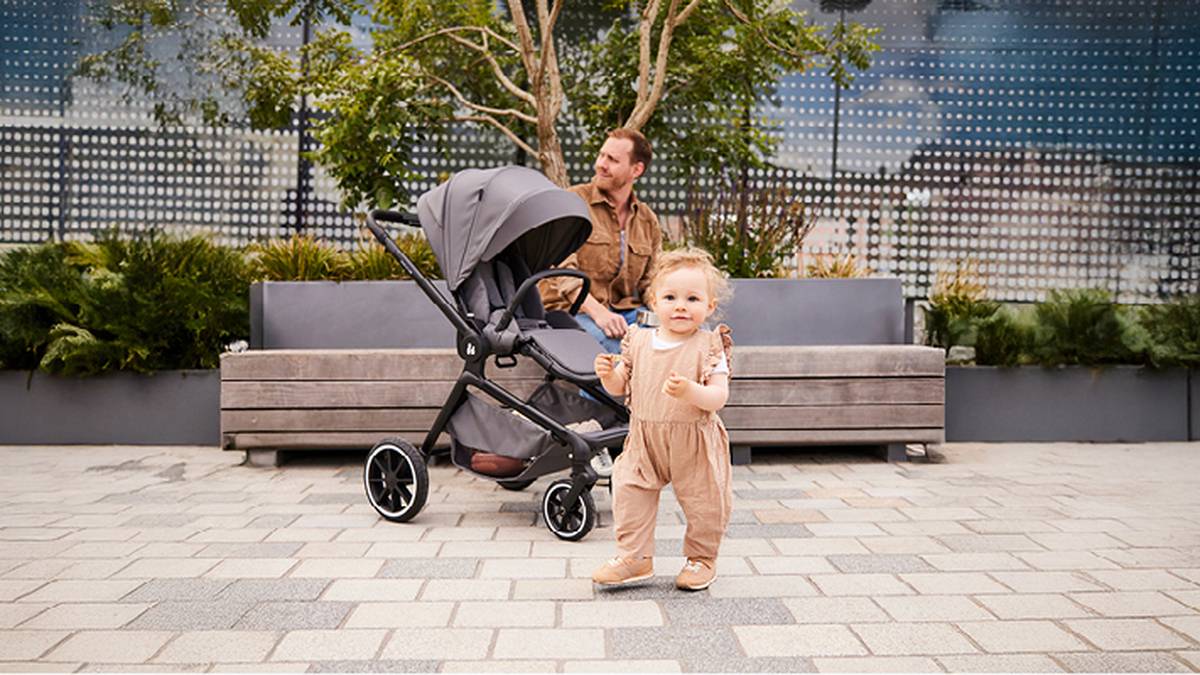 Ein Kleinkind in beigem Outfit läuft auf einem gepflasterten Platz, während ein Mann auf einer Bank hinter einem Kinderwagen unter Bäumen sitzt.
