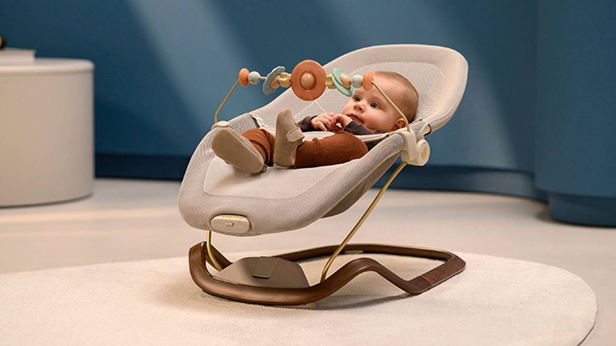 Ein Baby sitzt in einer modernen weißen Babywippe mit Spielzeugstange, die auf einem hellen Teppich in einem sanft beleuchteten Raum mit blauen Wänden steht.