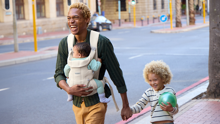 Ein Mann spaziert vergnügt durch eine Straße, hält die Hand eines Kindes und trägt ein Baby in einer Babytrage. Das Kind hält einen grünen Ball.