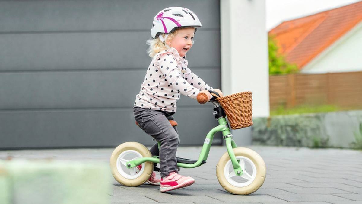 Ein kleines Kind mit Helm fährt auf einem kleinen grünen Laufrad mit Weidenkorb auf einem gepflasterten Weg neben modernen Häusern.