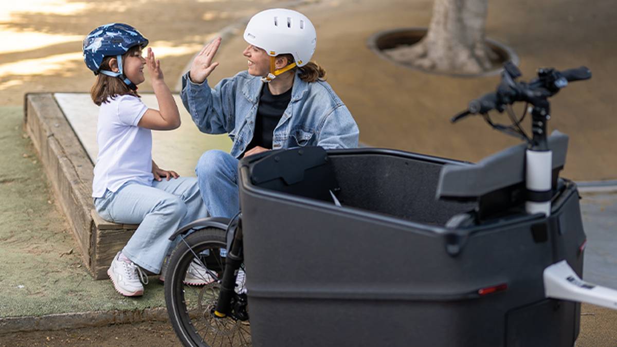 Eine Frau und ein Mädchen mit Helmen geben sich neben einem Lastenrad ein High-Five. Das Mädchen sitzt lächelnd auf einem Holzsims in einem Park.