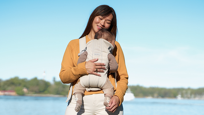 Eine Frau lächelt, während sie ein Baby in einer Trage hält. Sie steht im Freien in der Nähe eines Gewässers und trägt einen gelben Pullover unter einem klaren, blauen Himmel.