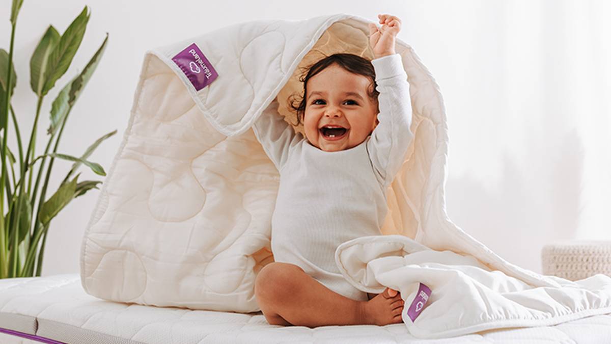 Ein fröhliches Baby in weißer Kleidung sitzt unter einer Steppdecke und streckt die Arme aus, im Hintergrund ist eine Pflanze zu sehen.