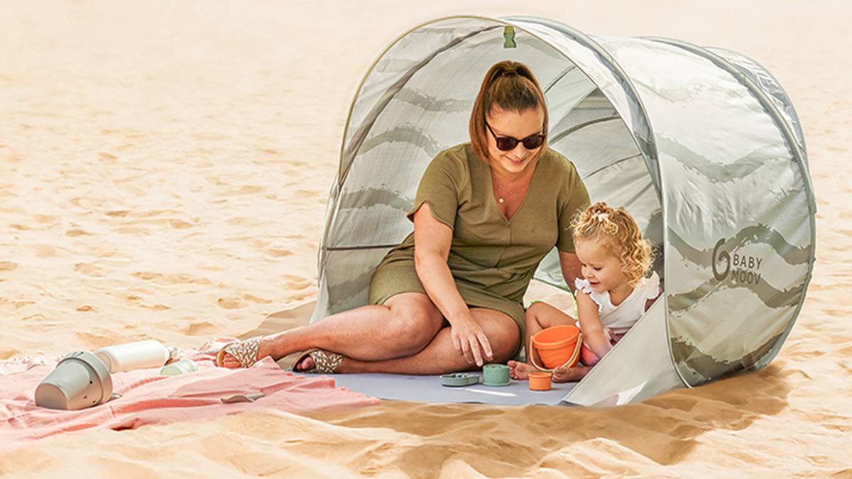 Eine Frau und ein Kind spielen mit Sandspielzeug unter einem kleinen Zelt an einem Sandstrand; daneben liegen ein zusammengerolltes Handtuch und Sonnencreme.