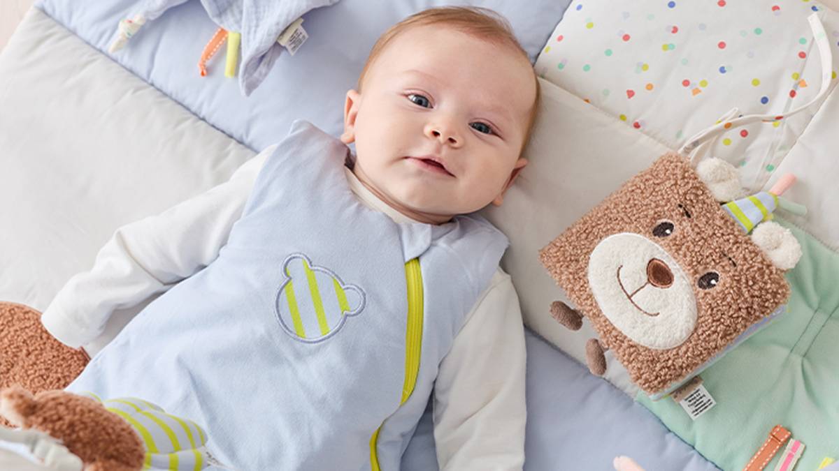 Ein Baby in einem hellblauen Outfit mit Bärenmotiv liegt auf einer weichen, bunten Decke neben einem lächelnden Bärenplüschtier.