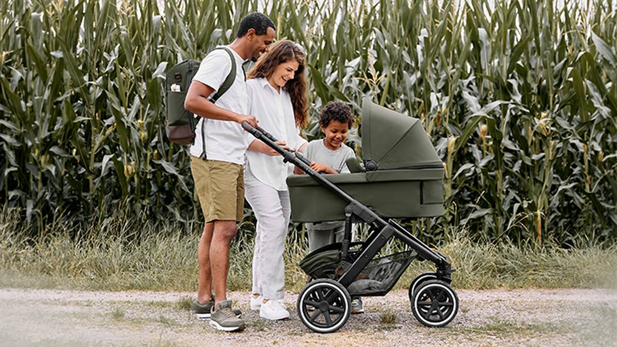Eine Paar mit einem Kind schauen zusammen das Baby im Kinderwagen an. Im Hintergrund ist ein Maisfeld