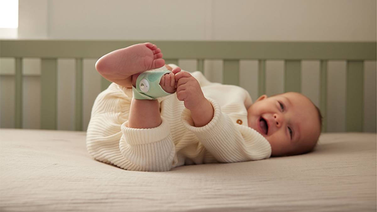 Ein Baby in einem weißen Outfit liegt auf einer Kinderbettmatratze und hält spielerisch seine Füße. An einem Fuß trägt das Baby ein kleines Gerät.