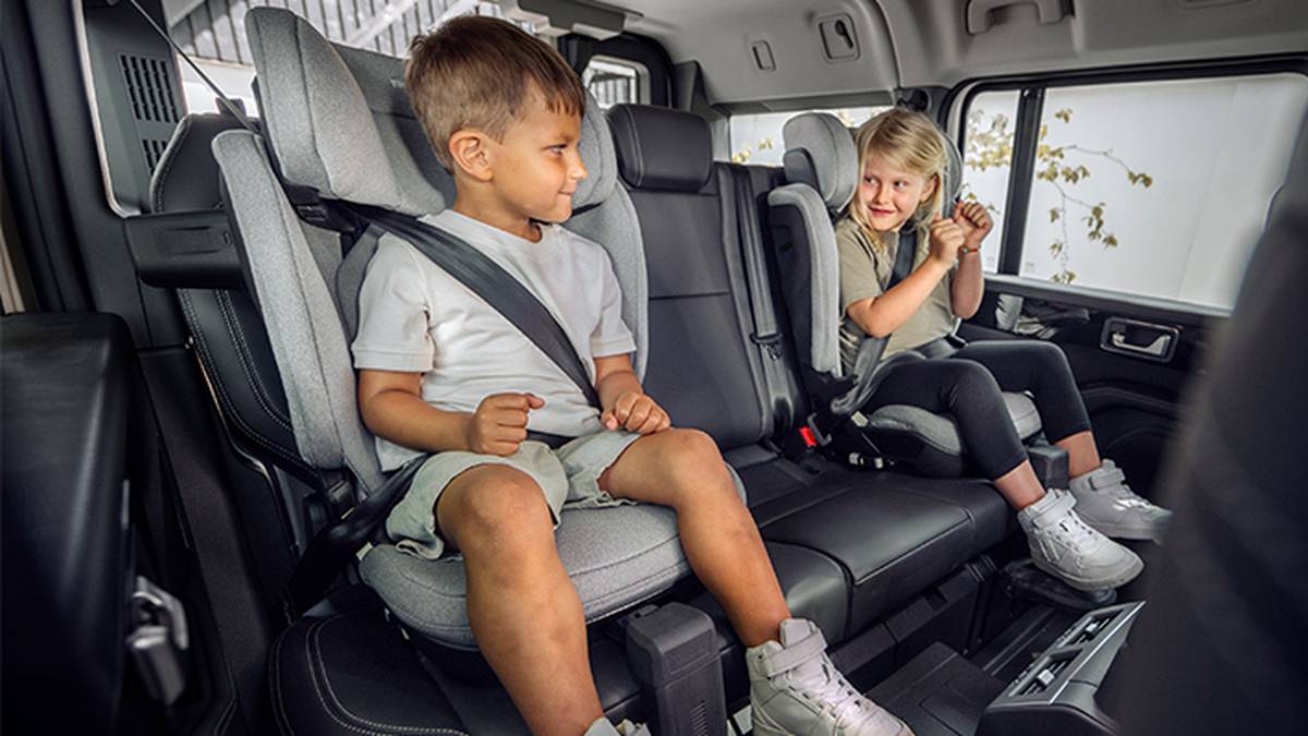Zwei lächelnde Kinder sitzen angeschnallt in ihren Kindersitzen in einem Auto. Der Innenraum ist modern und hell.