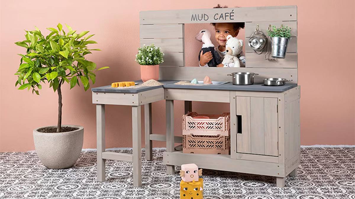 Ein Kind spielt in einer Spielküche, die einem „Matschcafé“ nachempfunden ist, mit Töpfen und Stofftieren. In der Nähe steht auf einem gemusterten Boden eine Topfpflanze.