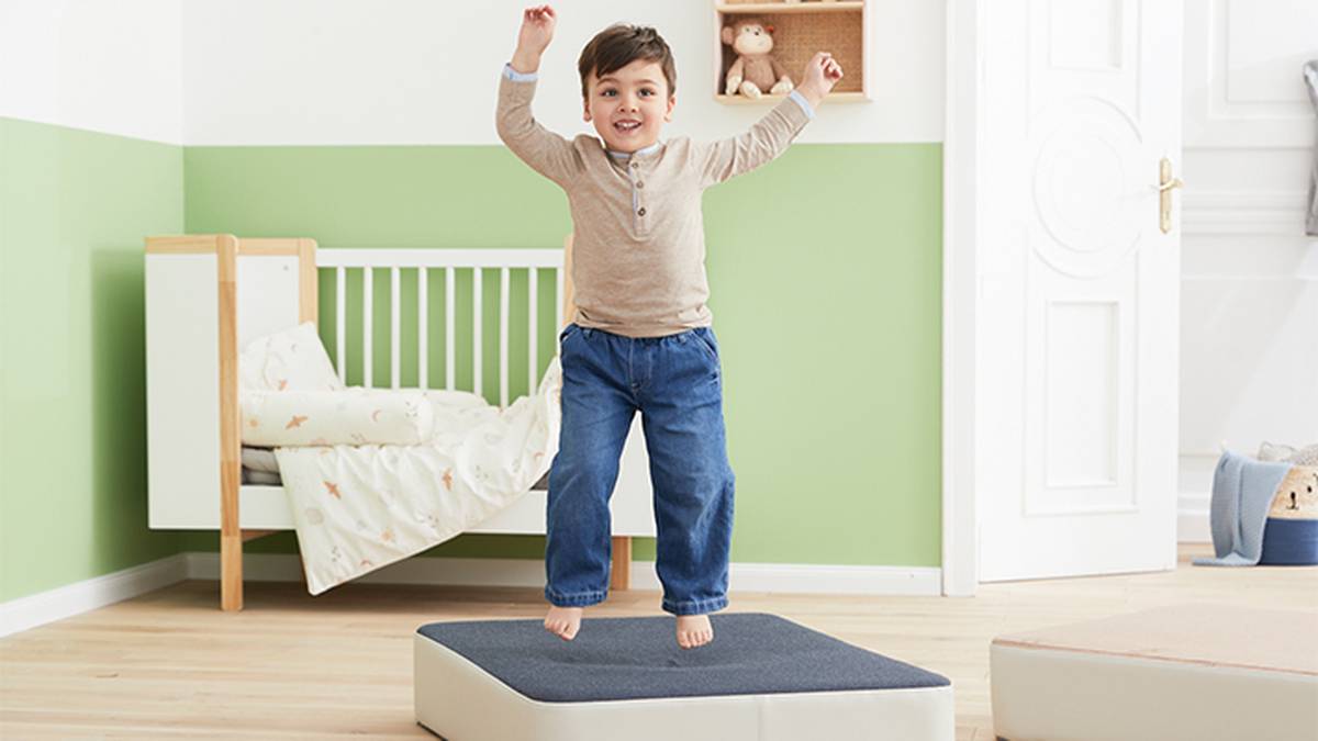 Ein Kind springt vergnügt auf einer gepolsterten Plattform in einem hellen, ordentlichen Zimmer; im Hintergrund sind ein Kinderbett und Spielzeug zu sehen.