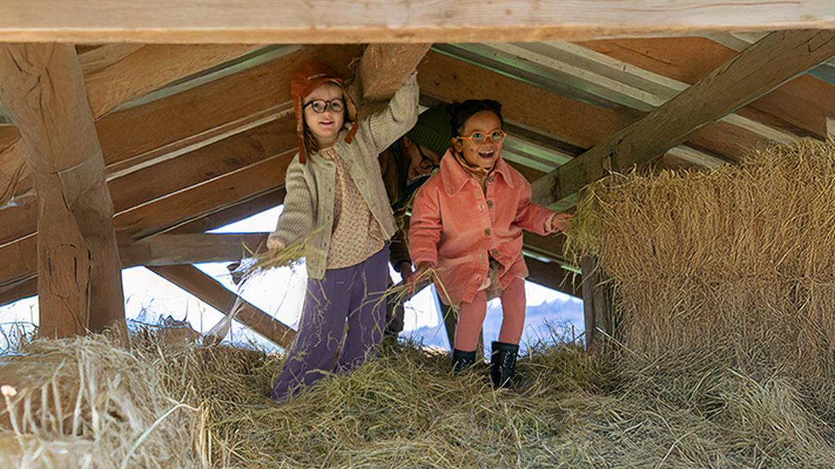 Zwei Kinder mit Brillen und warmer Kleidung spielen lächelnd und genießen ihre gemeinsame Zeit auf einem mit Stroh gefüllten Heuboden.