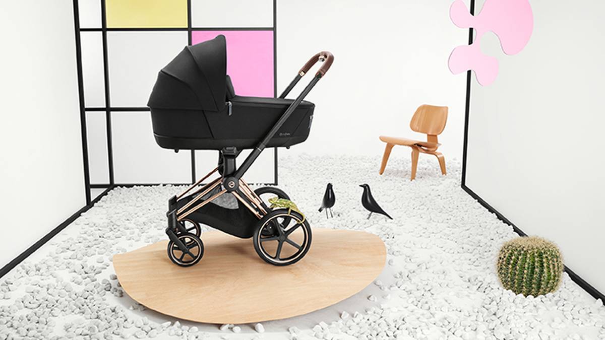 Ein schwarzer Kinderwagen mit goldenen Akzenten auf einer Holzplattform, umgeben von weißen Kieselsteinen, schwarzen Vögeln, einem Kaktus und einem Holzstuhl.